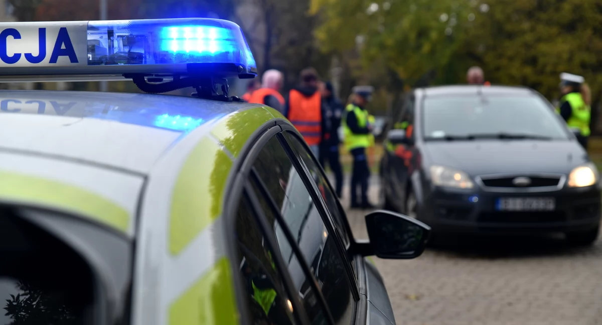 Zdjęcie policyjnego radiowozu na tle samochodu i policjantów/ podlaska.policja.gov.pl
