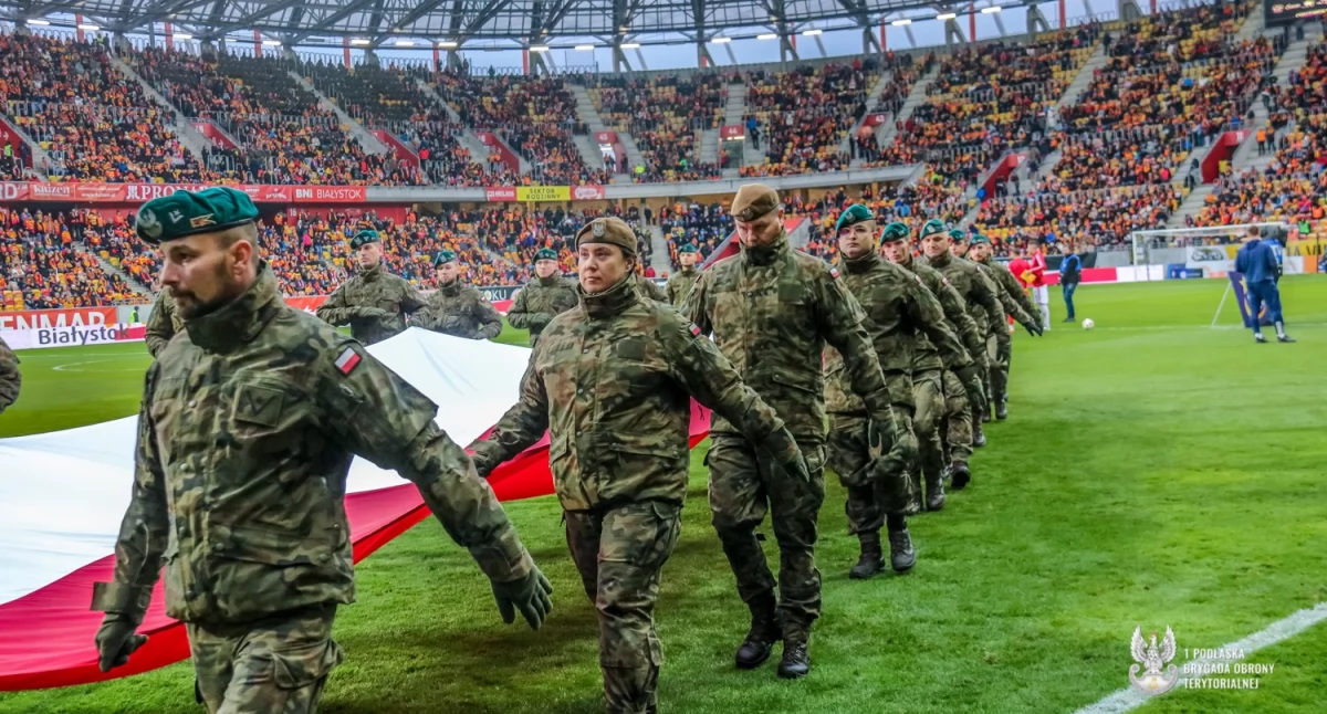 Zdjęcie terytorialsów niosących flagę narodową po płycie boiska stadionu miejskiego w Białymstoku/ 1PBOT